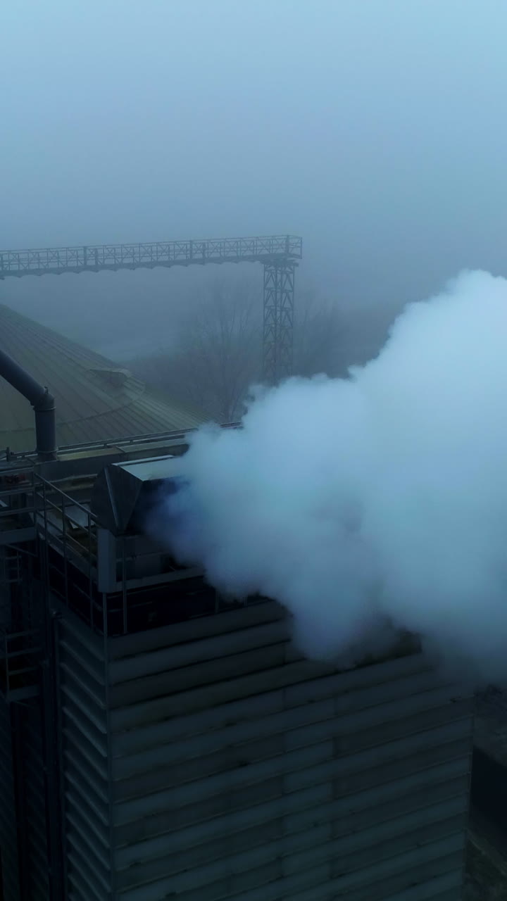 Powerful agribusiness plant for grain storage and processing. Metal tanks and high building with pipes producing thick smoke. Foggy backdrop. Top view. Vertical video