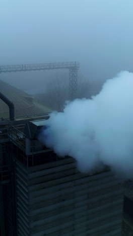 powerful agribusiness plant for grain storage and processing. metal tanks and high building with pipes producing thick smoke. foggy backdrop. top view. vertical video