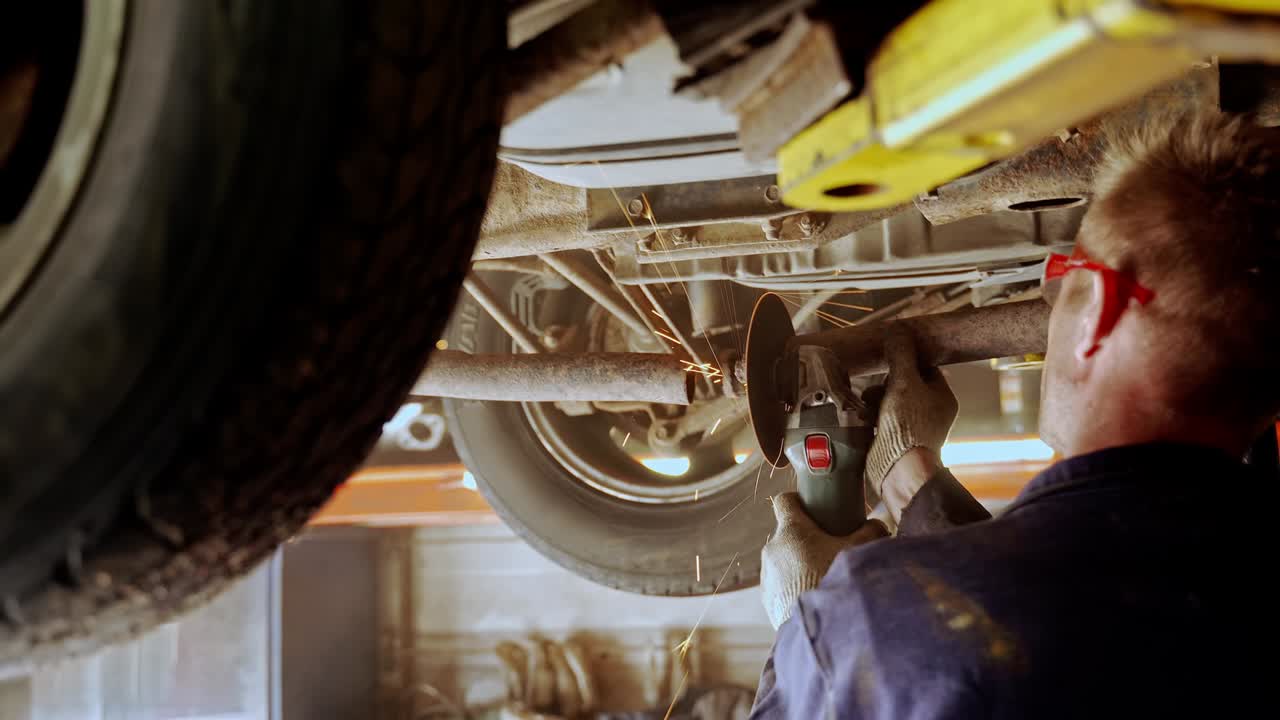 un hombre corta un tubo de silenciador con una molienda de ángulo. un trabajador en uniforme bajo la parte inferior de un coche en un ascensor
