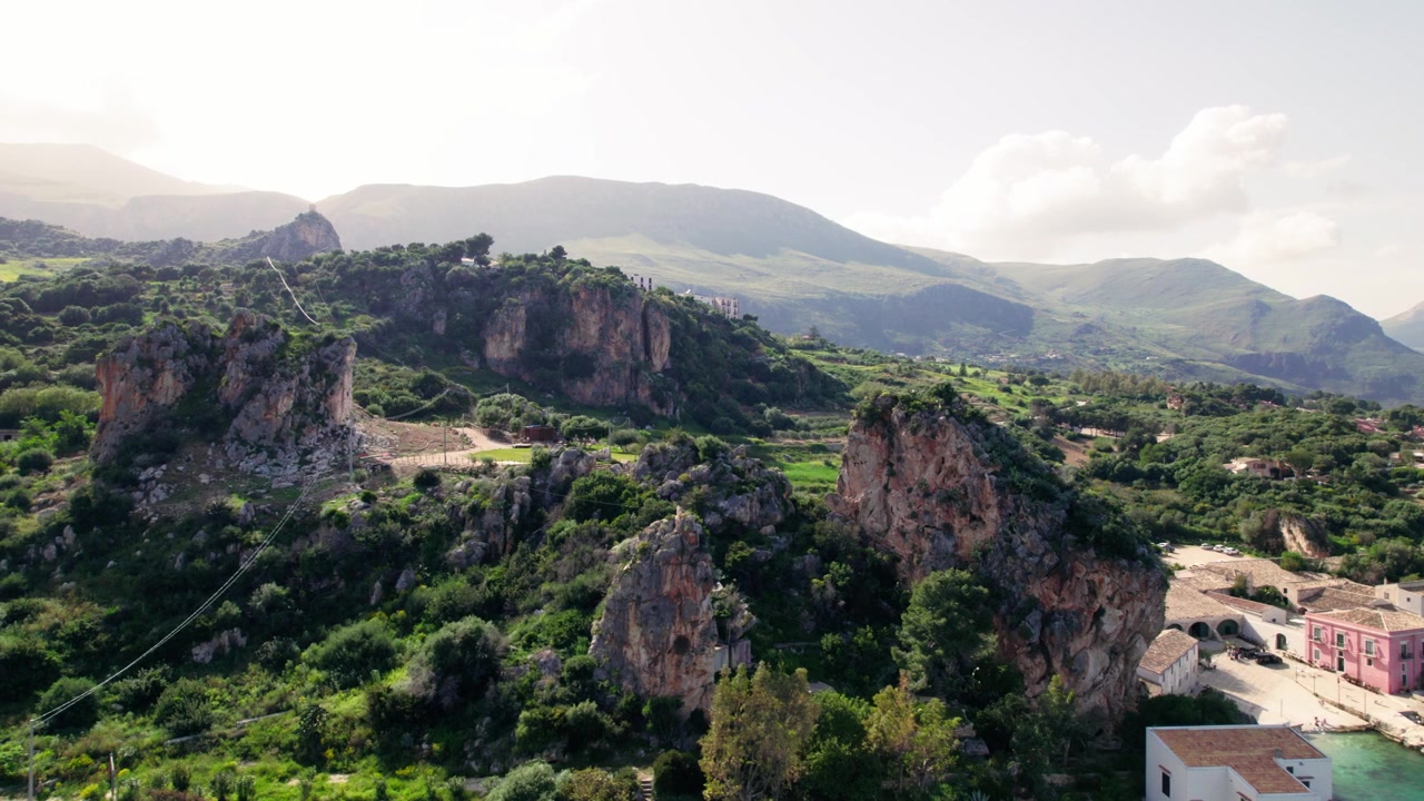 Una cresta calcarea frastagliata, coperta da vegetazione spontanea, si affaccia su una valle fertile nella campagna siciliana, incorniciata da montagne distanti.