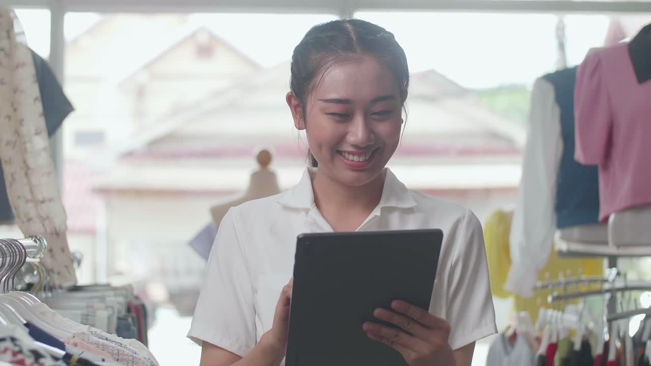 레일에 있는 재고를 확인하기 위해 디지털 태블릿을 사용하는 동안 카메라에 미소 짓는 옷 가게의 아시아 여성 소유자의 클로즈업