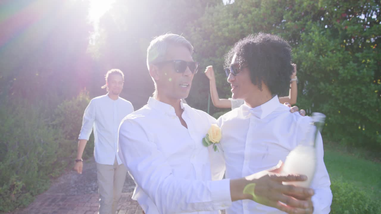 Gruppo eterogeneo che cammina su un sentiero, due uomini che si abbracciano mentre camminano, festeggiando, tenendo in mano una bottiglia e una spilla.