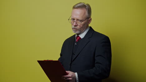 senior man in costume is writing. mature businessman in glasses holding a folder and thinking. portrait of an old professional entrepreneur on yellow background.