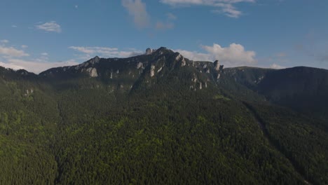 Aerial-view-showing-tall-mountains-peaks-covered-in-dense-coniferous-green-forests,-partly-cloudy-blue-sky