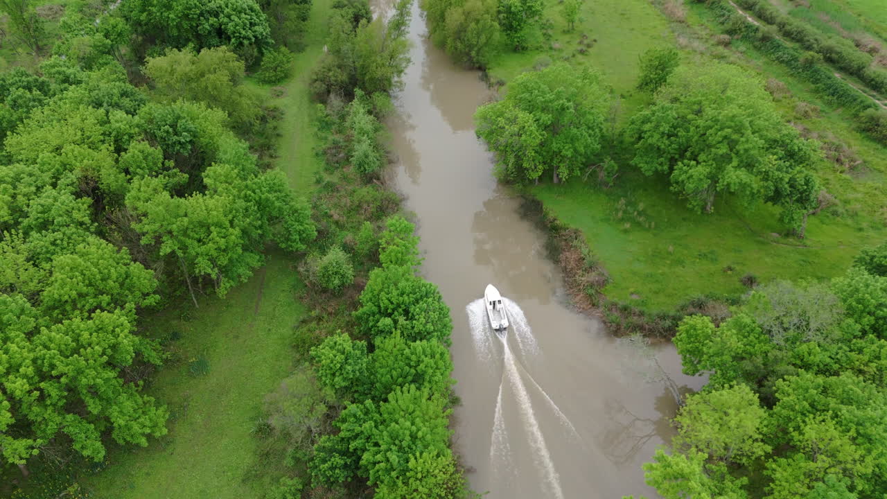 Drone track shot follows a small boat speeding along a narrow Delta del Paraná river, bordered by dense forest and wetland grasslands, muddy waters stirred by engine wake