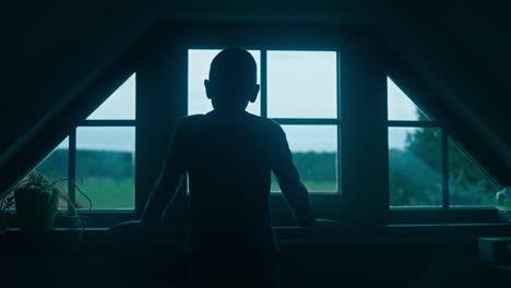 silhouette of a young boy child looking out the window at dusk in a dark room before he goes to bed