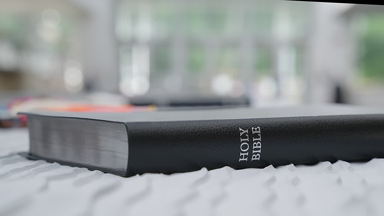 Black leather Holy Bible rests on white bedding with soft, natural morning window light