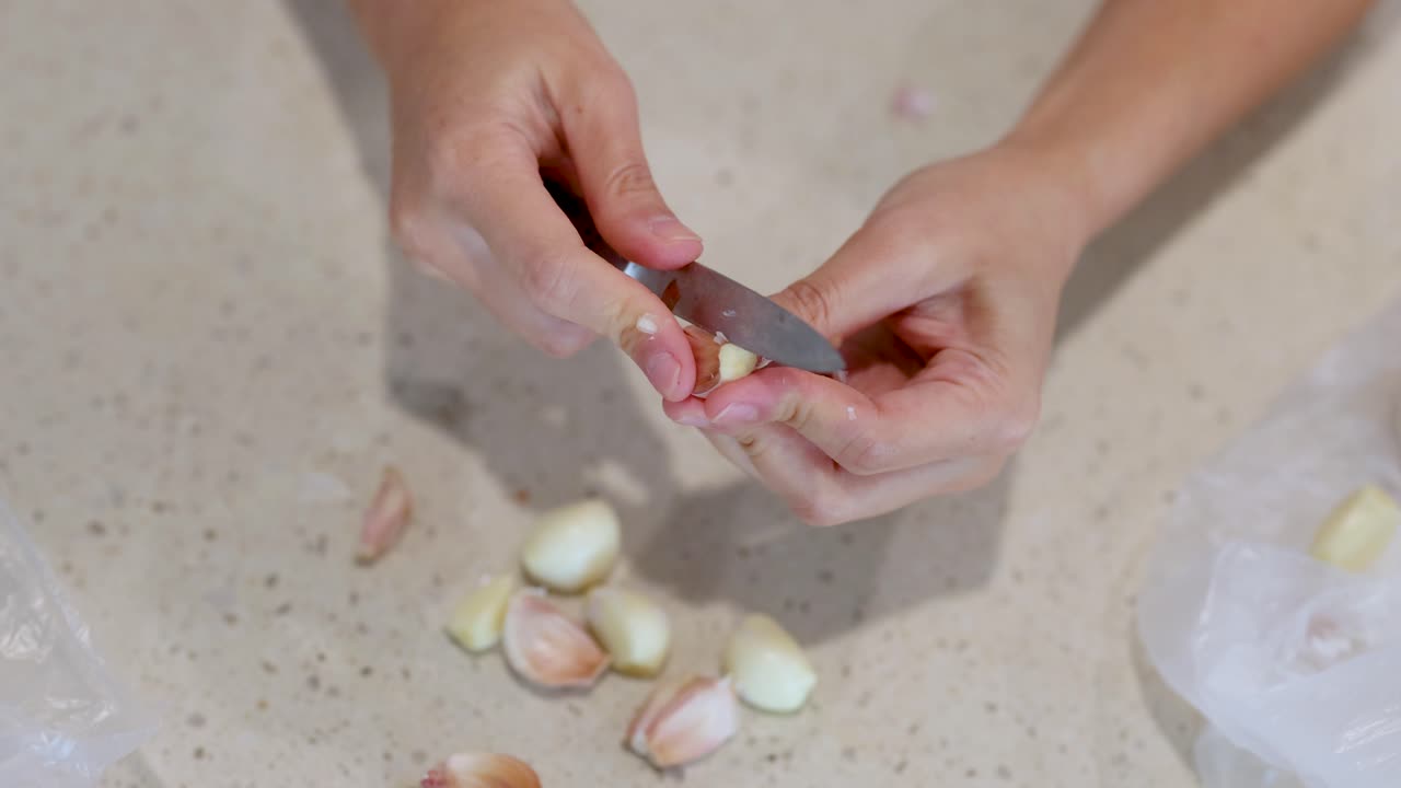 Close-up of hands peeling garlic cloves on a light kitchen countertop. Natural lighting, steady camera, focused on culinary preparation