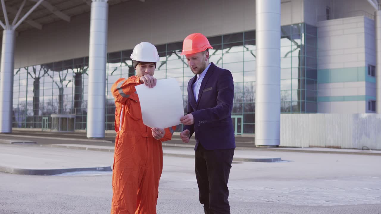 un joven arquitecto con un traje negro examinando el objeto de construcción con un trabajador de la construcción con uniforme y casco naranja. se encuentran en el objeto de construcción y se dan la mano.