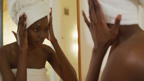 african american woman with towel touching her face and watching in mirror in bathroom