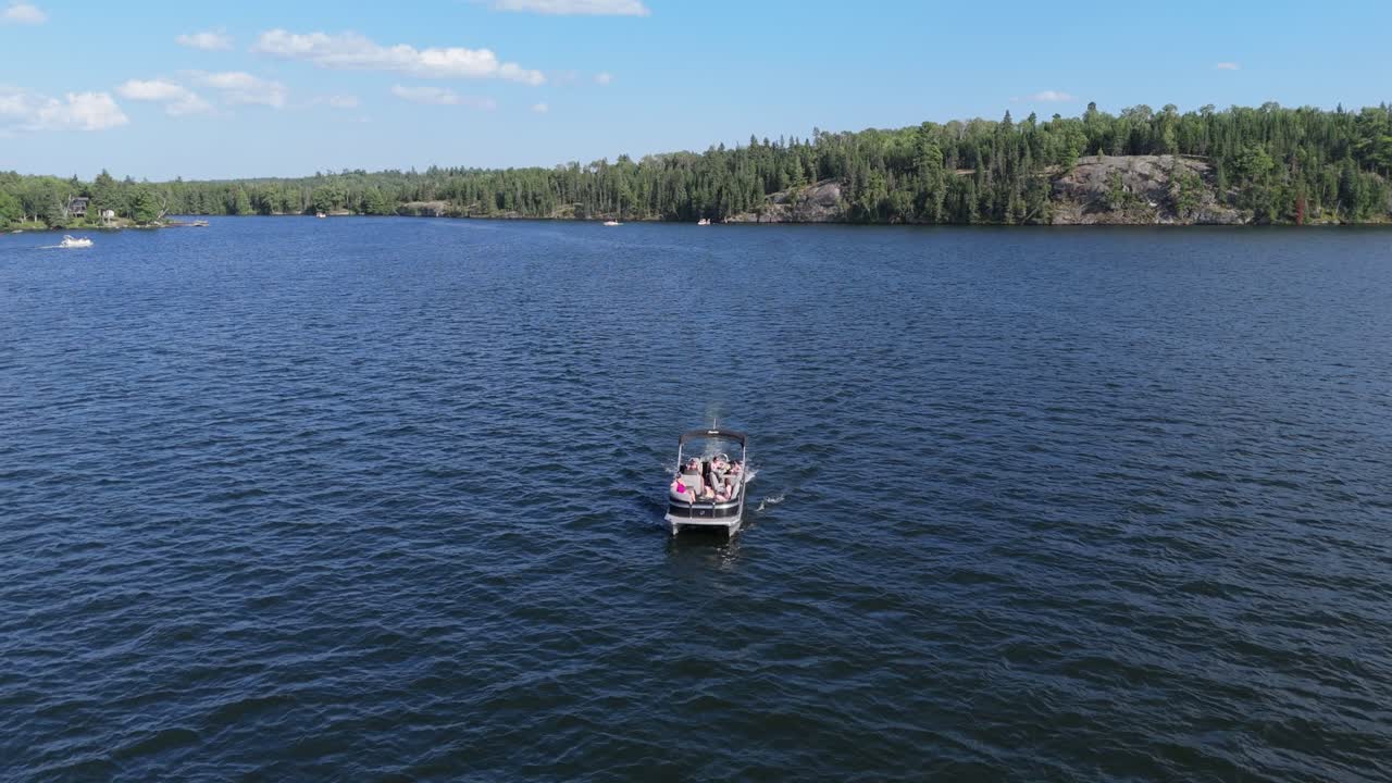 Aerial drone footage of a motorboat cruising across a blue lake toward the camera, with forest and rocky shoreline in the background