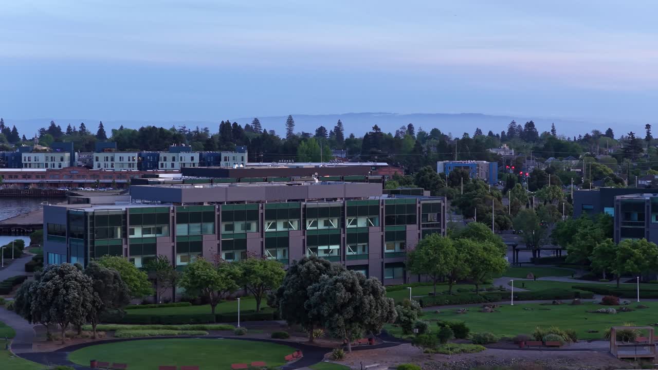 Vista panorámica orbital con dron de un moderno complejo de oficinas y el parque circundante en Alameda, California, capturada al anochecer con las luces de la ciudad empezando a encenderse y montañas visibles a lo lejos.