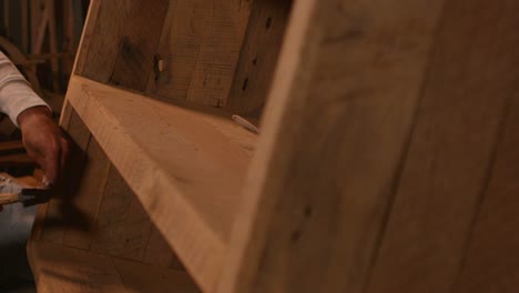 the camera pans left to reveal a woodworker hammering a nail into a bookshelf