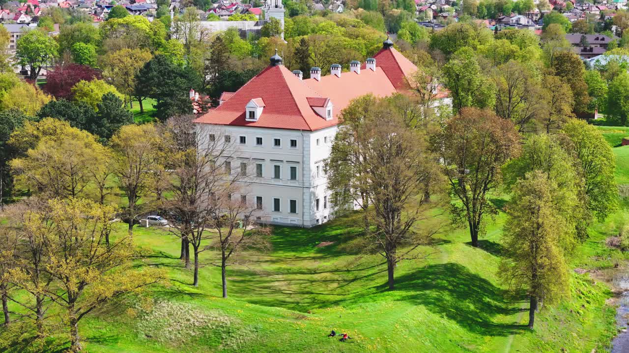 A historic white estate with a red roof stands on a green hill among tall trees in a European park, surrounded by blooming spring foliage and distant rooftops.