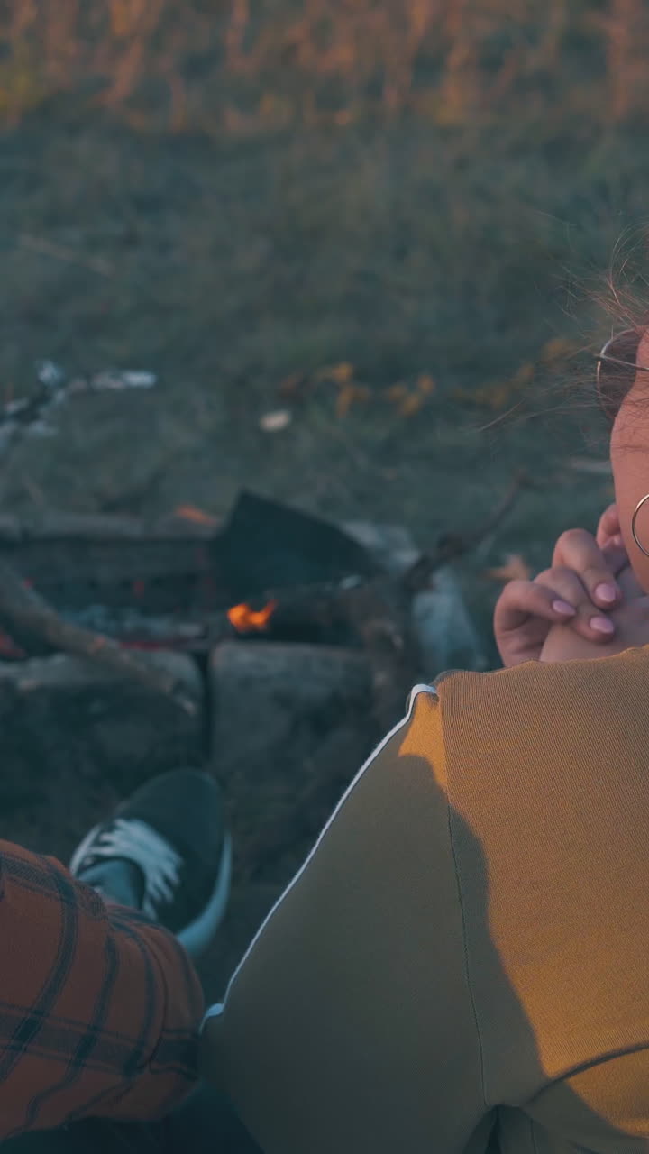 cheerful plump girl with glasses tells story to friends at burning bonfire on meadow in autumn evening backside view