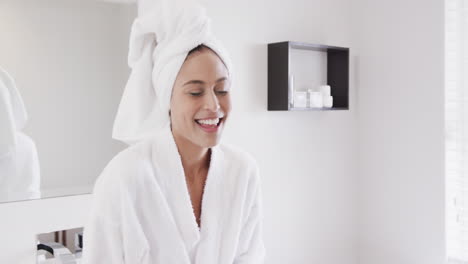 Retrato-De-Una-Mujer-Birracial-Feliz-Usando-Una-Toalla-En-La-Cabeza-Usando-Un-Teléfono-Inteligente-En-El-Baño,-Cámara-Lenta