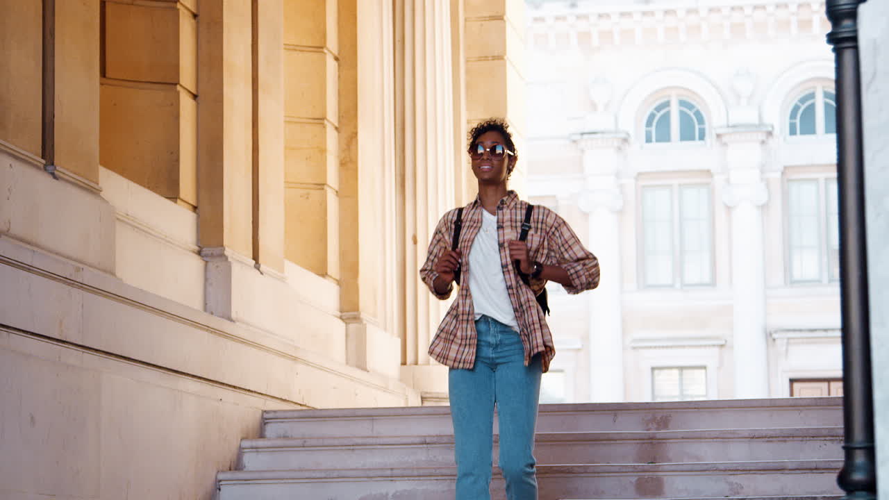 Young black woman wearing a plaid shirt and blue jeans walking down street stairs at the entrance of a historical building, full length