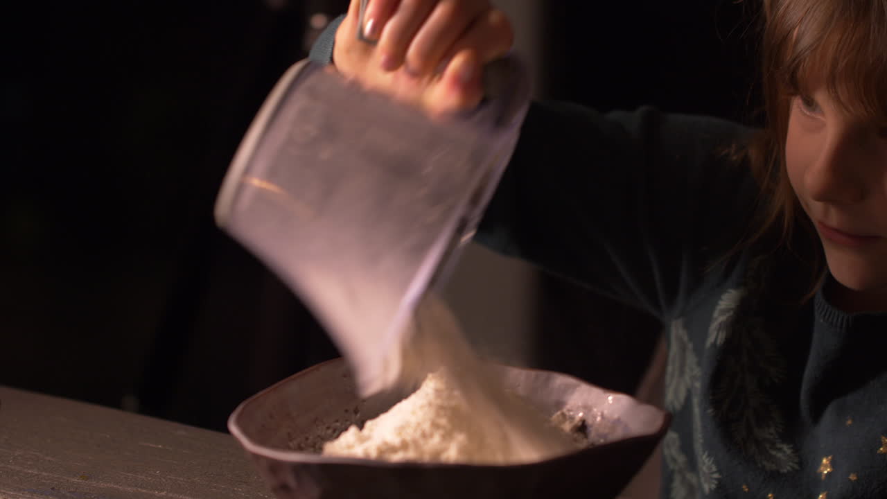 Adorable little girl pouring baking flour from measuring cup to bowl in kitchen