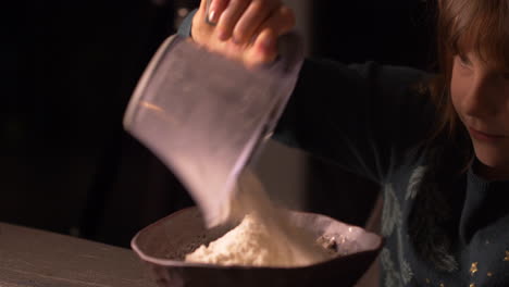 adorable little girl pouring baking flour from measuring cup to bowl in kitchen