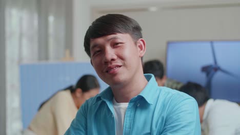 미소 짓는 아시아 남성 엔지니어가 동료들이 사무실에서 풍력 터빈에 대해 논의하는 동안 카메라에 팔을 고 있습니다.