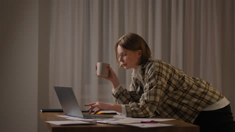 a young beautiful business woman works from home with a cup of coffee and looks into a laptop screen standing at the table. view mail and drink coffee. remote work from home