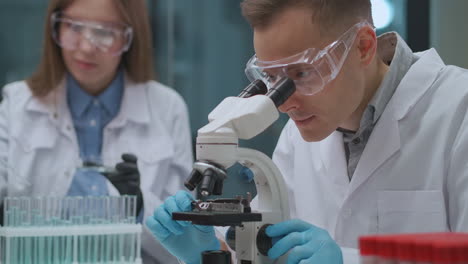 Un-Par-De-Científicos-Está-Trabajando-En-Un-Laboratorio-Criminalístico-El-Hombre-Está-Viendo-Evidencia-En-Un-Microscopio-La-Mujer-Está-Probando-Líquidos-En-Tubos-De-Ensayo-Práctica-Experta-Forense