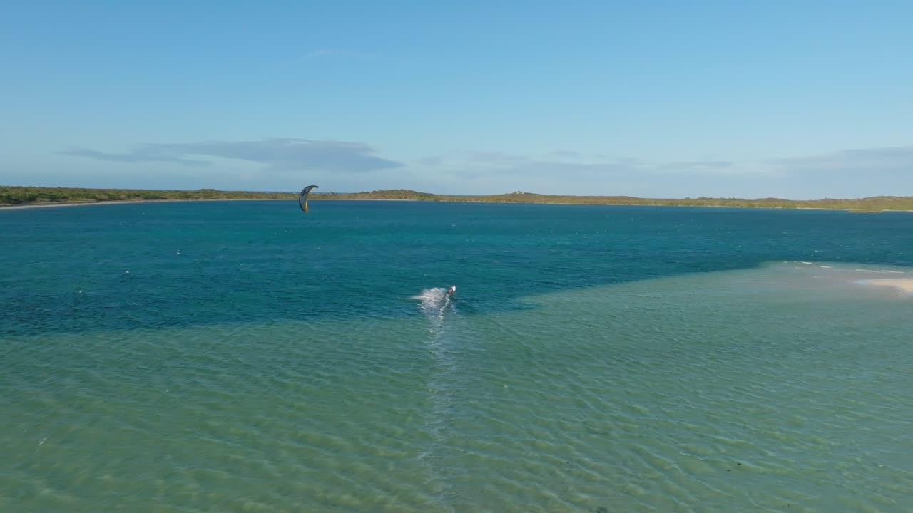 Drone follows a kitesurfer gliding from the beach across clear turquoise waters near a private island