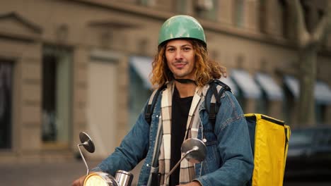 Portrait-of-a-happy-courier-guy-with-long-curly-hair-in-a-denim-jacket-who-sits-on-a-moped-and-looks-at-the-camera-along-with-his-big-yellow-food-delivery-bag-in-a-summer-city