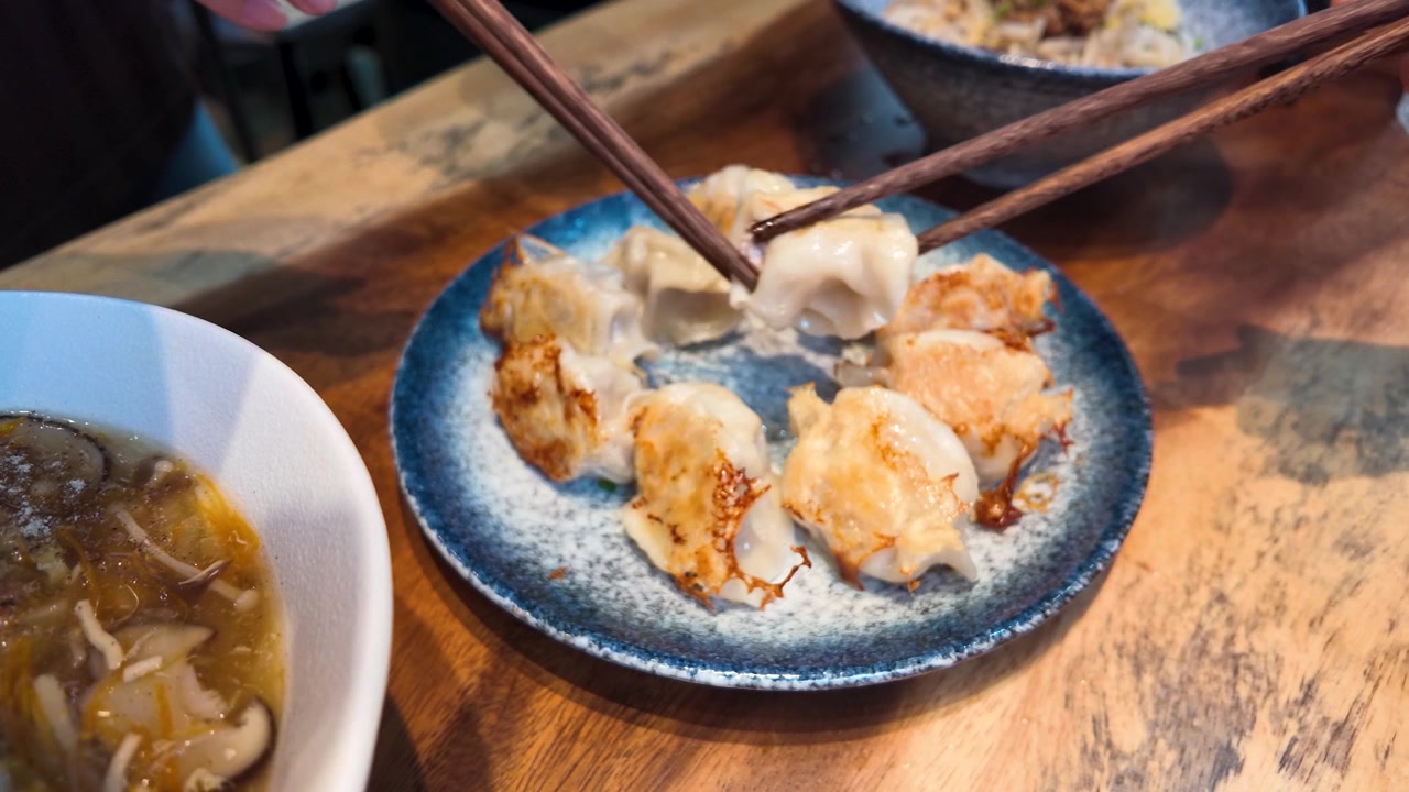 Close-up of wooden chopsticks picking up pan-fried dumplings from a blue ceramic plate in Bangkok.