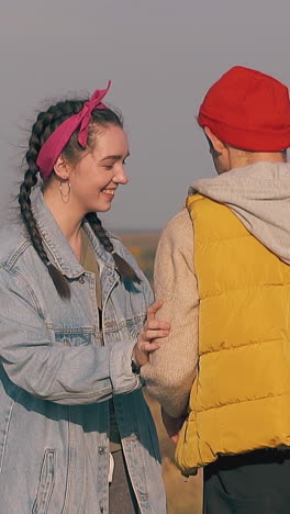 smiling girl in denim jacket gives guy with hangover red bottle with water in morning against river slow motion