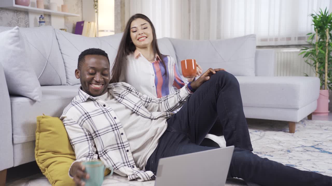 African man and caucasian woman working from laptop at home, drinking coffee. Loving multiethnic married couple.