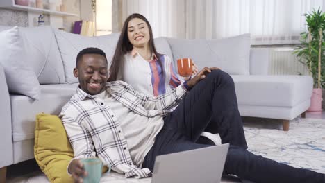 un hombre africano y una mujer caucásica trabajando desde una computadora portátil en casa, bebiendo café. una pareja casada multiétnica enamorada.