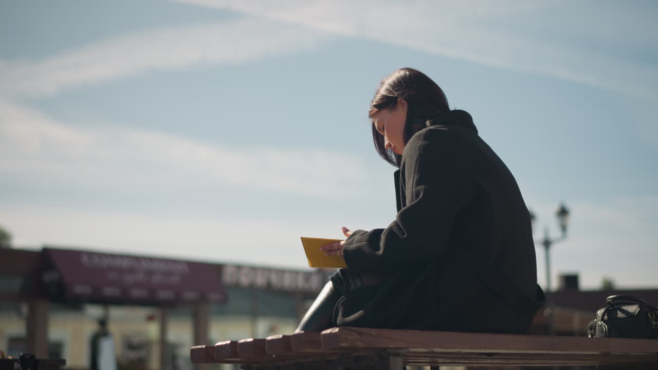 Woman in black coat sitting outdoors on bench, reading book with yellow-edged pages visible, background features urban buildings, lamp posts, and bright sunlight in a public space