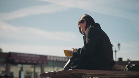 woman in black coat sitting outdoors on bench, reading book with yellow-edged pages visible, background features urban buildings, lamp posts, and bright sunlight in a public space