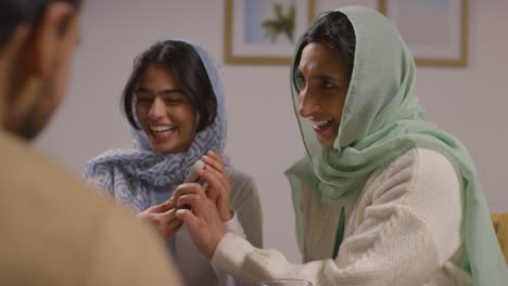 Muslim-Mother-And-Daughter-Laughing-At-Mobile-Phone-Sitting-Around-Muslim-Family-Table-At-Home