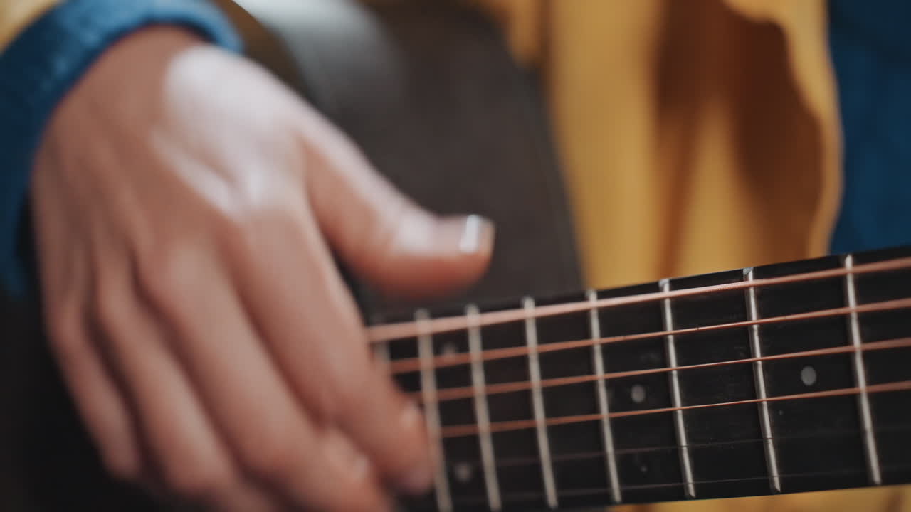 Mani in primo piano che strimpellano una chitarra acustica, campeggiatore che indossa una giacca gialla, movimento di fingerpicking sulle tastiere di legno, atmosfera calda e soffusa, sottile bagliore del fuoco da campo, suono intimo e rustico per video di viaggio