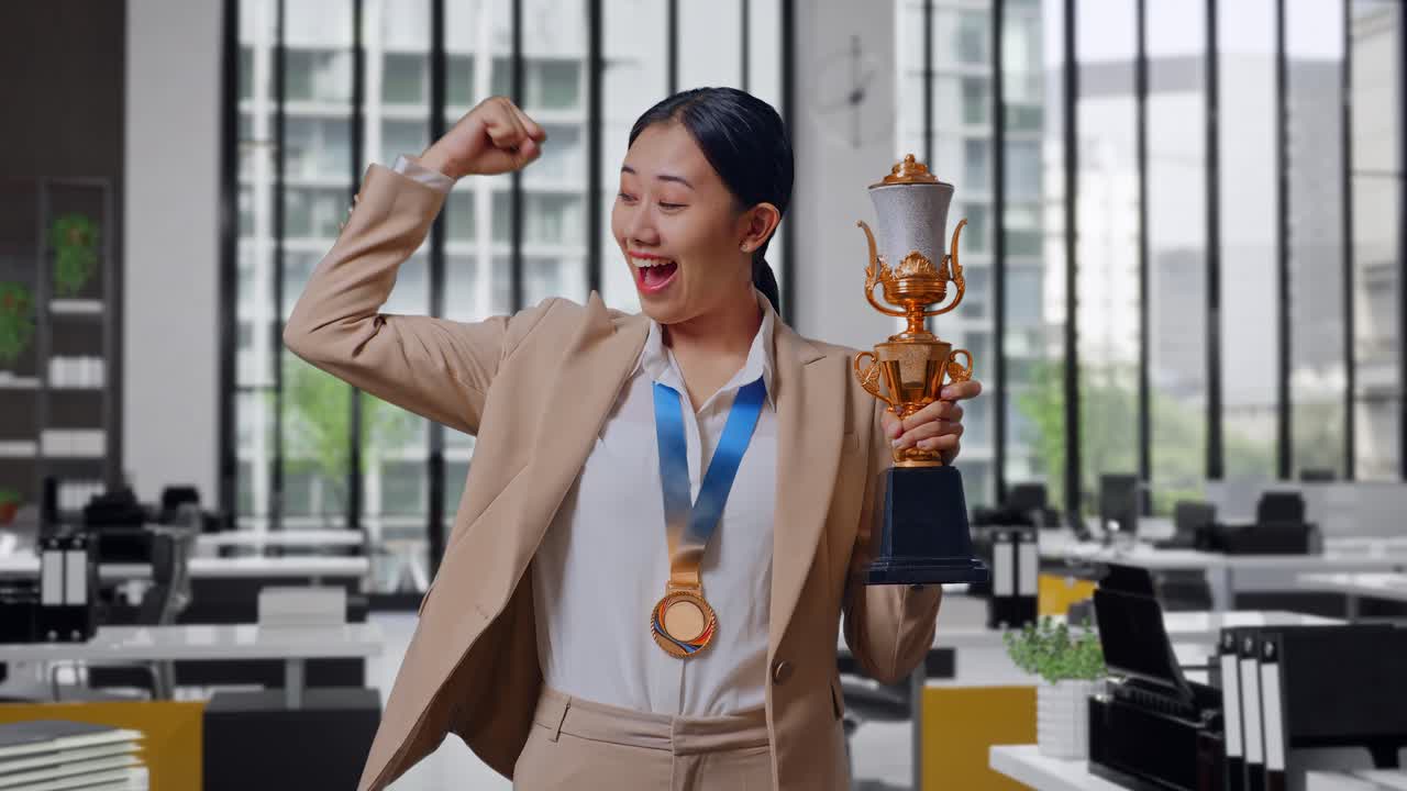 Asian Business Woman In A Suit With A Gold Medal And Trophy Flexing Her Bicep And Smiling To Camera In The Office