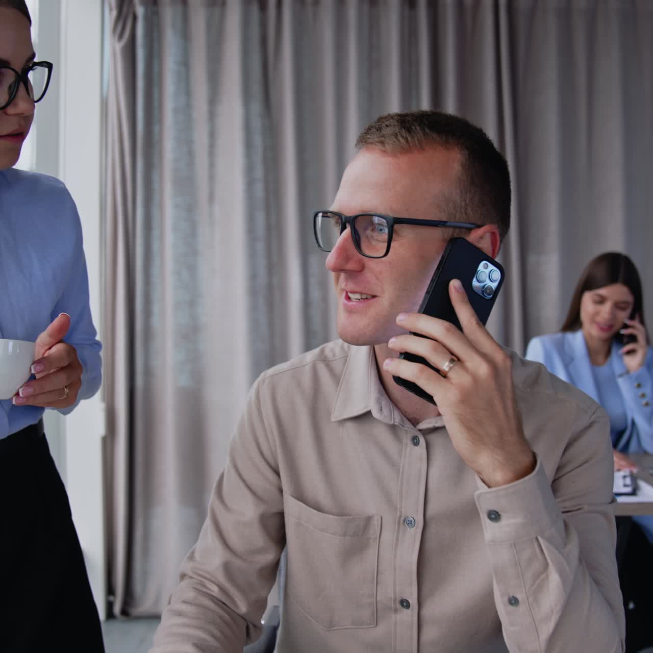 Un impiegato seduto alla scrivania inizia una conversazione telefonica. Una donna con una camicia blu e una tazza di caffè in mano pone delle domande, guardando il portatile.