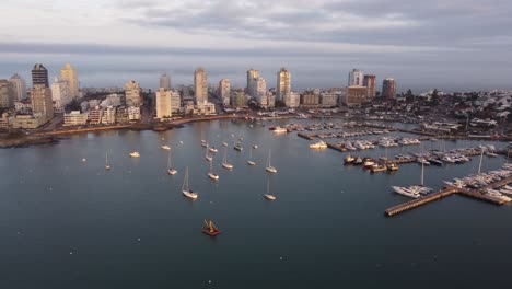 Luftaufnahme-Des-Hafens-Mit-Verankerten-Segelbooten-Vor-Der-Stadt-Punta-Del-Este-Bei-Sonnenuntergang---Rückwärtsflug