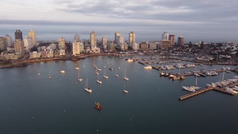 aerial view of port with anchored sailing boats in front of punta del este city at sunset - backwards flight