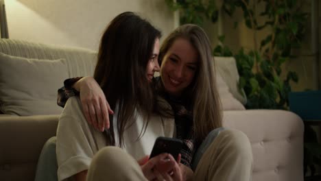 a smiling lgbt couple of girls are using smartphone while sitting on the floor together