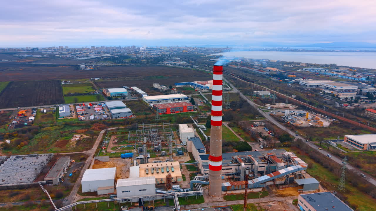 Sítio industrial urbano com chaminé. Fumaça sobe de uma chaminé alta em um sítio industrial perto do horizonte de uma cidade e campos abertos.