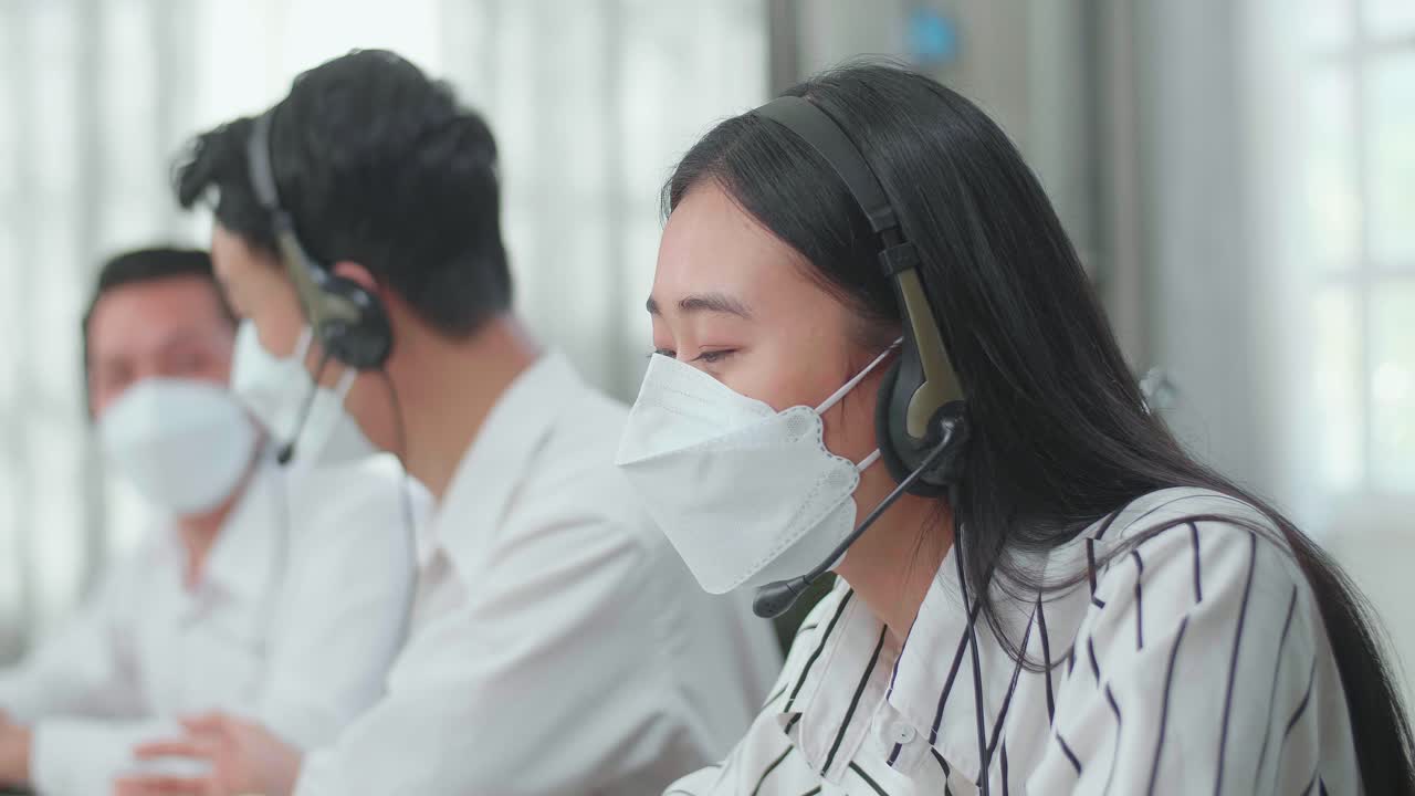Close Up Of A Woman Of Three Asian Call Center Agents Wearing Headset And Mask Speaking To Customer On The Call While Two Of Her Colleagues Are Talking During Working At The Office