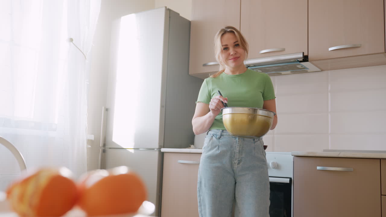 Žena v zelené košili a džínách drží mísu a míchá mouku v prosvětlené kuchyni, usmívá se, zatímco pomeranče jsou rozmazané v popředí poblíž prosluněného okna se záclonou