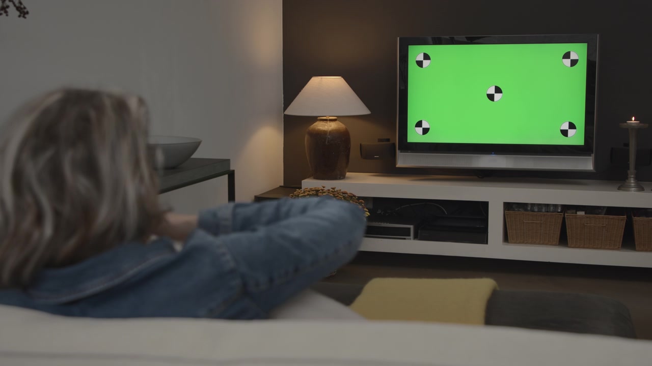 Inquadratura ampia di una giovane donna che guarda la TV in un soggiorno moderno con opzioni di green screen chroma key per effetti visivi.