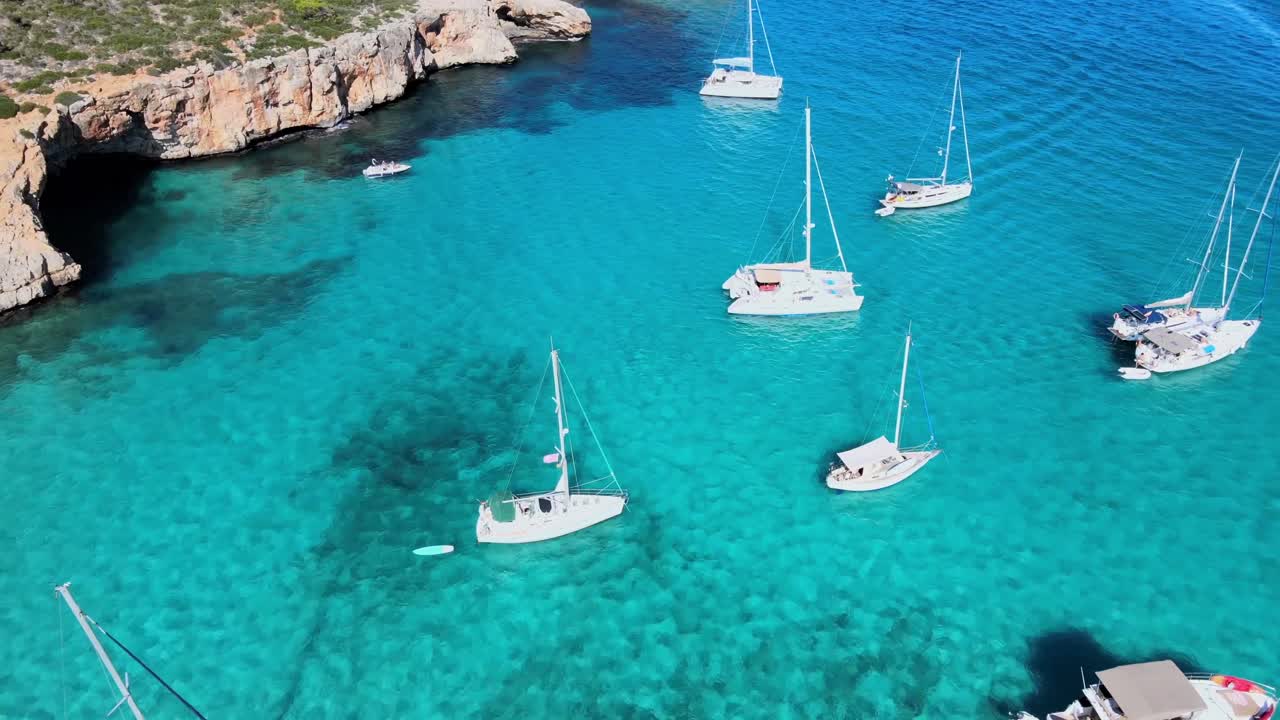 Oplev den rolige skønhed ved Cala Varques, Mallorca, med talrige sejlbåde forankret i krystalklart vand. Nyd de omkringliggende klipper og frodige grønne områder under en klar blå himmel