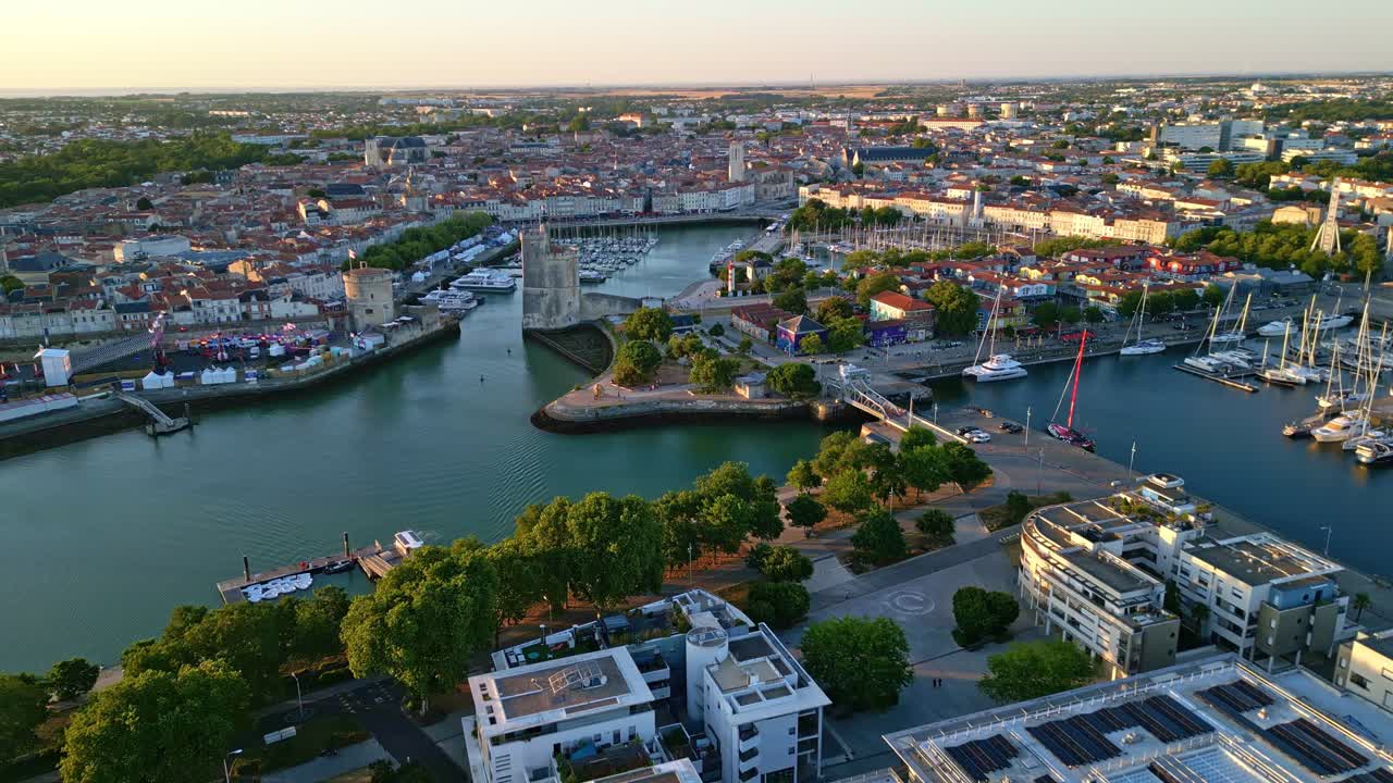 Backward drone shot over La Rochelle harbor at sunset, revealing the Lantern Tower, boats, colorful houses, green trees and cityscape - France