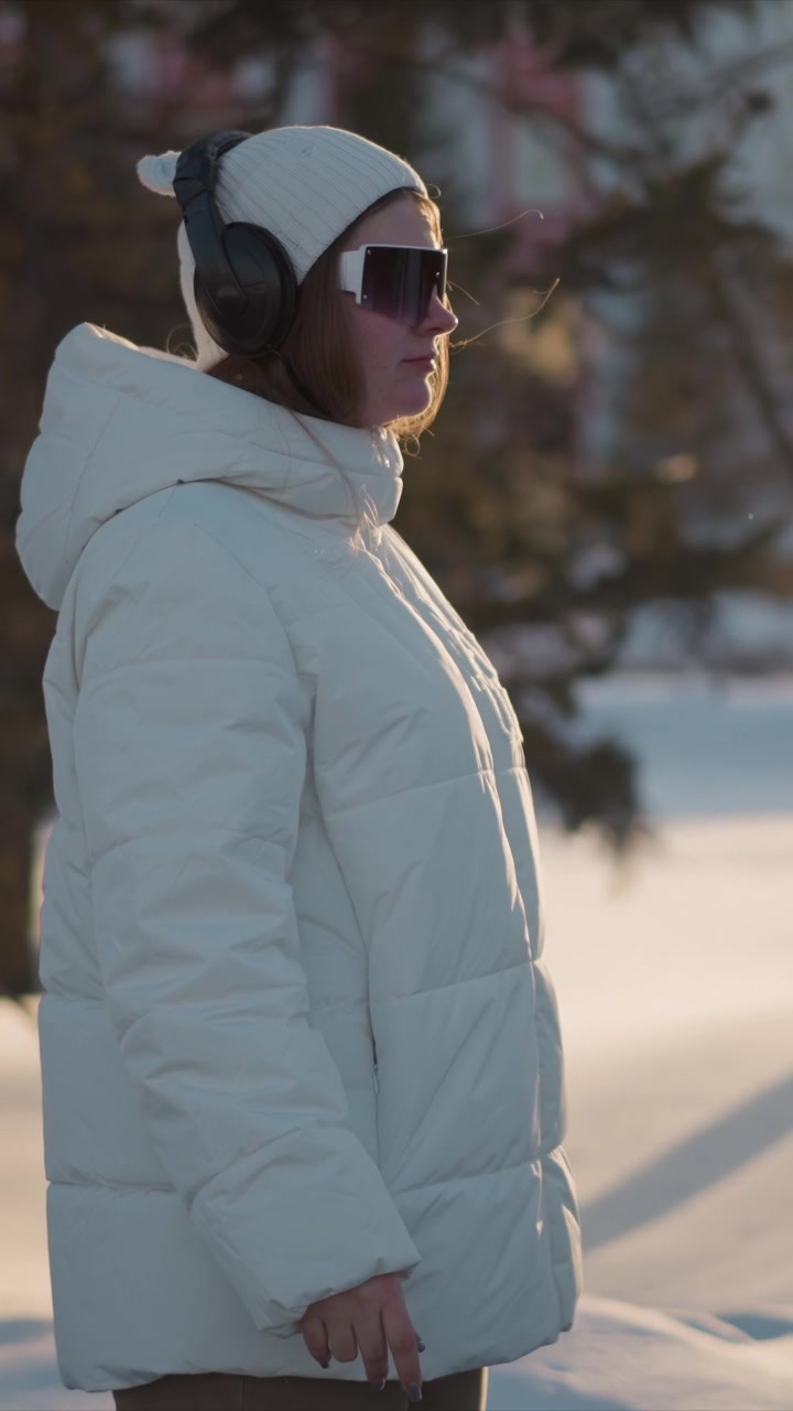 Woman wearing white puffer jacket knit hat with earflaps and headphones walks outdoors in snowy environment. Woman holds sunglasses or object near her face while background features trees