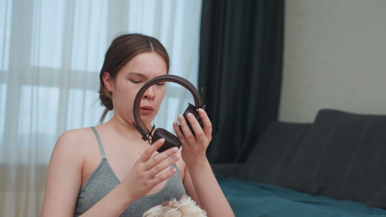 Donna bianca incinta che spacchetta delle cuffie in camera da letto, espressione curiosa mentre prova i padiglioni auricolari e ispeziona il cavo, seduta sul letto con un peluche e luce diurna soffusa, canotta casual e maternità rilassata