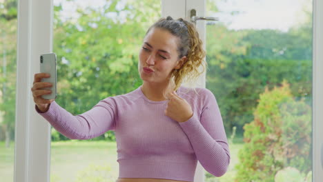 young woman sitting on yoga mat at home after exercise posing for selfie on mobile phone